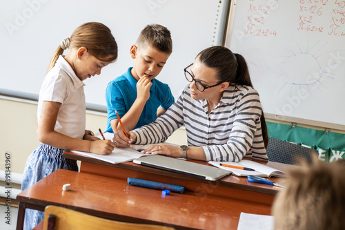 In the classroom's cozy setting, the caring female teacher diligently checks her elementary school students' homework, providing guidance and support to nurture their academic growth and success.