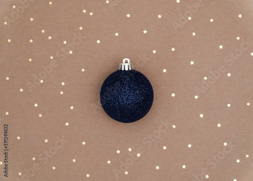 A classic navy blue glitter Christmas bauble in the centre of the frame. Gold sparkles in bokeh style in random pattern on backdrop of plain neutral brown paper. Copy space festive background.