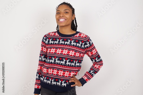 Studio shot of cheerful Young beautiful African American woman wearing Christmas sweater against white wall keeps hand on hip, smiles broadly.