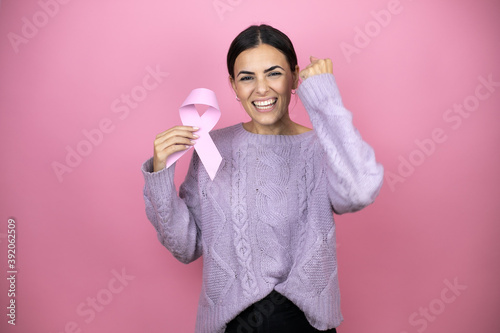 Beautiful woman wearing a casual violet sweater over pink background holding brest cancer ribbon screaming proud and celebrating victory and success very excited, cheering emotion