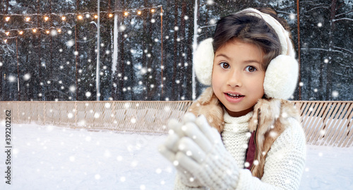 christmas, season and children concept - happy little girl wearing earmuffs over winter ice skating rink background