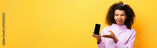 excited african american woman pointing with hand at smartphone with blank screen on yellow, banner
