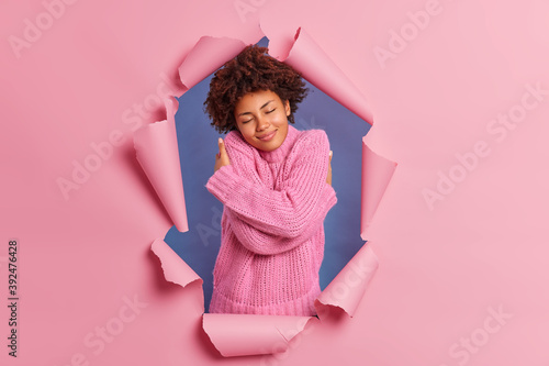 Photo of pleased African American woman embraces herself has high self esteem dressed in soft sweater closes eyes poses through ripped background in torn hole has dreamy expression imagines something
