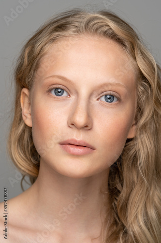 Close up of young woman face with blue eyes, curly natural blonde hair and eyebrows, has no makeup, looking at camera. Girl with perfect fresh clean skin over studio grey background. Youth concept. 