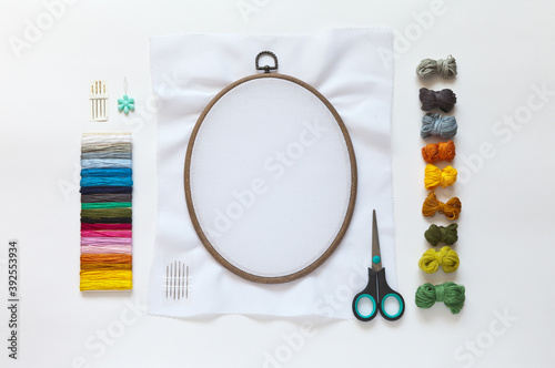 Accessories for embroidery: needles, set of colored mouline threads, needle threader, scissors and cotton canvas on the oval hoop for stitch or cross-stitch embroidery. Copy space, flat lay, top view