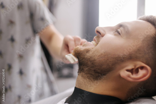 Professional hairdresser uses a hair clipper for fringing beard for a handsome man in barbershop. side view