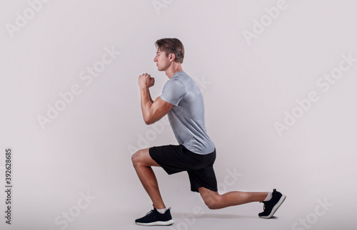 Side view of young fit guy in sportswear doing lunge on light studio background