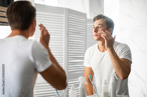 Handsome guy looking in the mirror applying cream on face