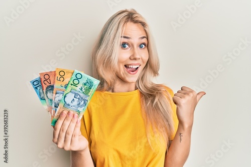 Beautiful caucasian blonde girl holding australian dollars pointing thumb up to the side smiling happy with open mouth