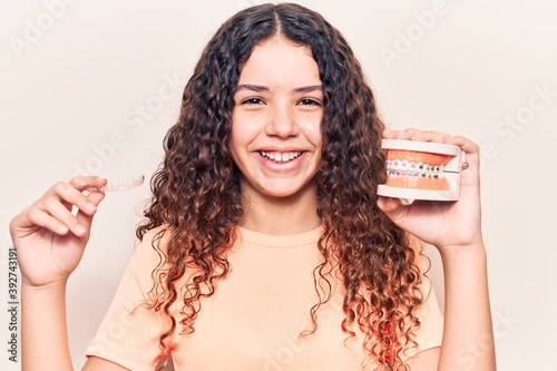 Adorable latin teenager smiling happy. Standing with smile on face holding denture with bracket and aligner over isolated white background