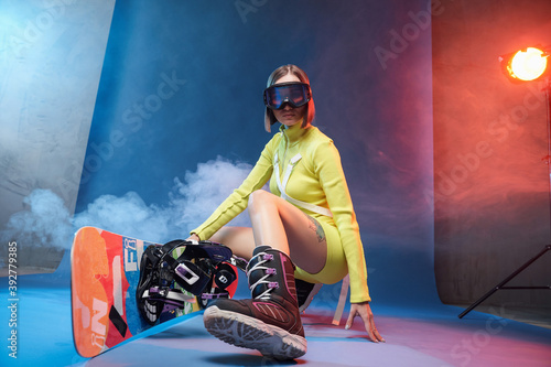 Cheerful young sportswoman in yellow sportswear with ski glasses and short haircut poses with snowboard in colored background with smoke.