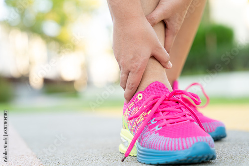 Female fatty runner athlete leg injury and pain. Hands grab painful ankle while running in the park.