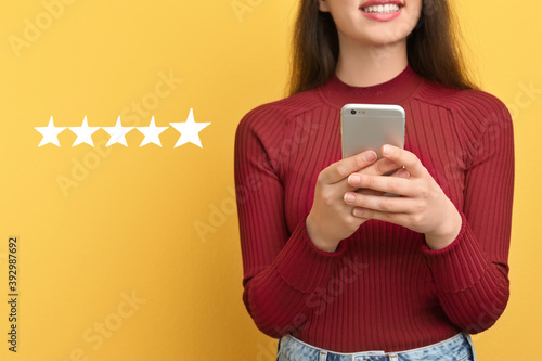 Woman leaving review online via smartphone on yellow background with five stars, closeup. Service or product feedback