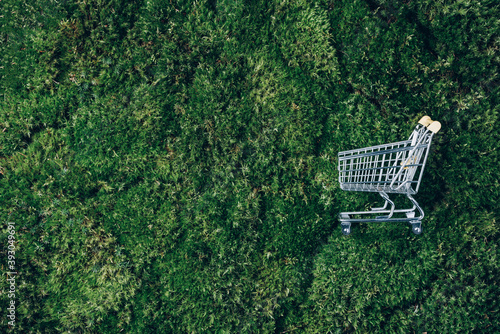 Shopping cart on green grass, moss background. Top view. Minimalism style. Creative design. Shop trolley. Sale, discount, shopaholism, ecology concept. Sustainable lifestyle, conscious consumption