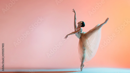 Young slim ballerina in light long dress is dancing on colored background with backlight