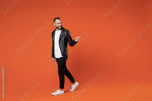 Full length side view of smiling young bearded man in basic white t-shirt black leather jacket standing pointing index finger aside on mock up copy space isolated on orange background studio portrait.