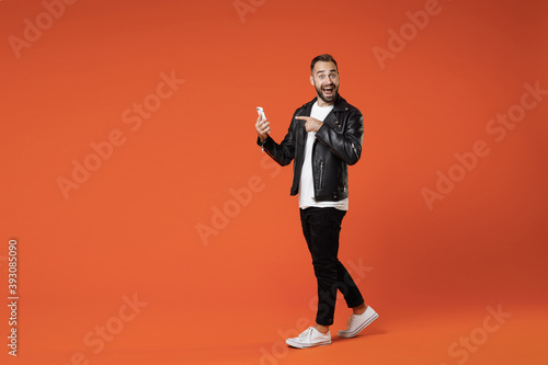 Full length side view of shocked young bearded man in basic white t-shirt black leather jacket standing pointing index finger on mobile cell phone isolated on orange colour background studio portrait.