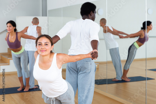 Positive young hispaic woman practicing couple yoga positions with african american partner during training in fitness center