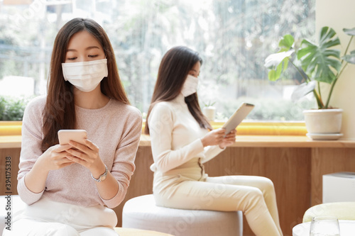 Two young Asian women wear face mask using smart phone and tablet while sitting at Public place. New normal lifestyle and social distance in city concept