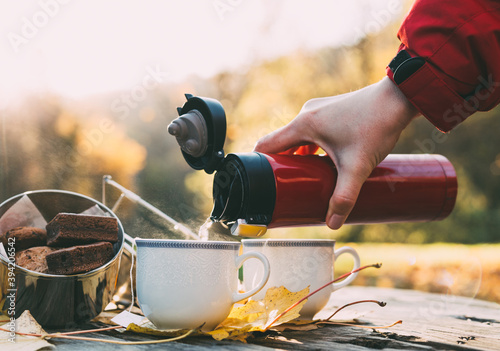 Early morning picnic in forest in late autumn. Cookies and hot tea woman pouring from thermos bottle on wooden table.