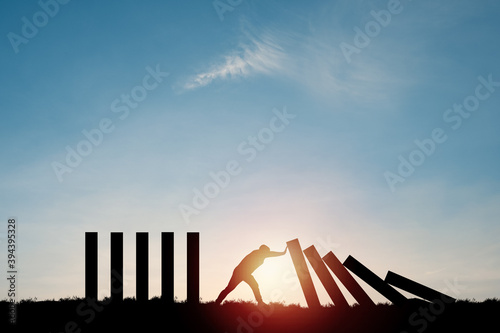 Silhouette man pushing rectangle block which falling to stop dominos others rectangle standing with blue sky. Risk and crisis management concept.