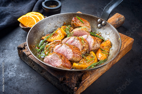 Traditional fried pork filet medaillons in with caramelized orange slices and herbs offered as close-up in a rustic wrought iron skillet