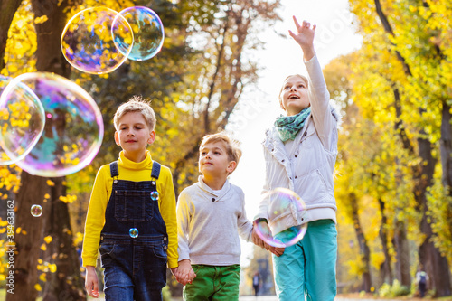 Three funny children girl and boys playing with soap bubbles, someone blows children catch bubbles. Beautiful natural landscape, aututmn park on background, happy childhood.