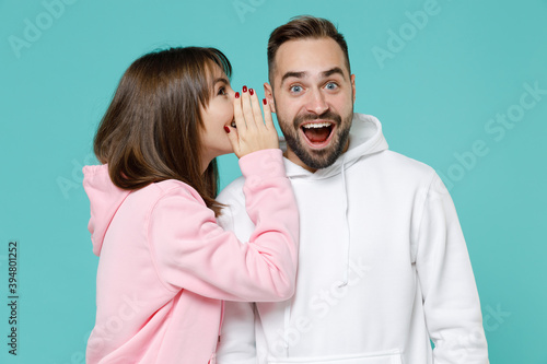 Excited young couple two friends man woman 20s wearing white pink casual hoodie standing whispering secret behind hand, sharing news isolated on blue turquoise colour wall background studio portrait.