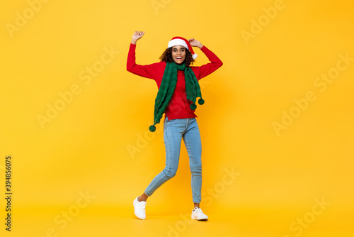 Fun portrait of happy African American woman in Christmas attire smiling and raising hands on isolated yellow studio background