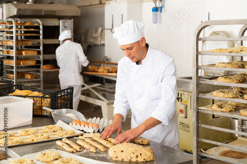 Working at bakery, male baker kneading dough and shaping baguettes on steel countertop in industrial kitchen