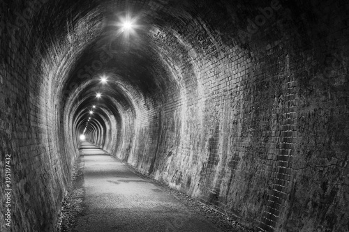An old tunnel with brick walls, lit up with electric lighting. Black and white. Photographed at the former Karangahake Gorge rail tunnel, New Zealand, opened in 1905