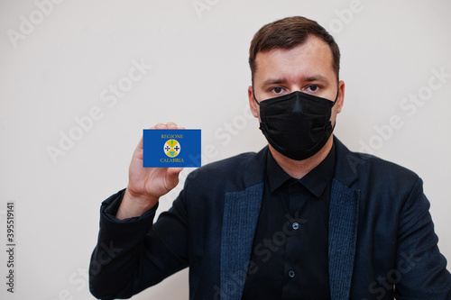 Italian man wear black formal and protect face mask, hold Calabria flag card isolated on white background. Italy regions coronavirus Covid concept.