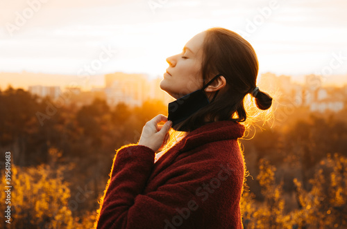 Side view of young woman taking the face mask off for a gulp of fresh air backlit by orange sunset light