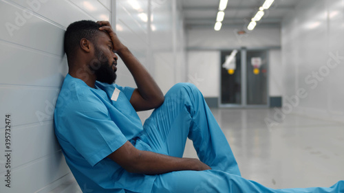Stressed tired young afro surgeon sitting on floor in hospital corridor after difficult surgery