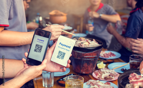 Hand holding smartphone to scan Qr code payment tag with blurry grilled pork and meat on the stove, Thai food buffet payment tag. QR code payment concept to many friends at a party in a restaurant.