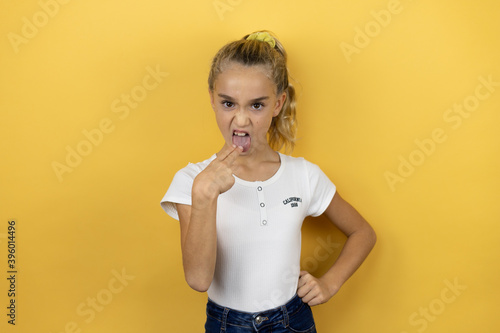 Young beautiful child girl standing over isolated yellow background disgusted with her hand inside her mouth