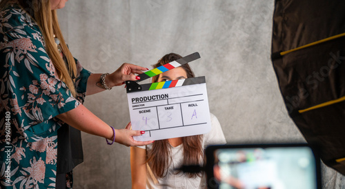 Operator holding clapperboard during the production of short film inside a studio with young actress on stage. Focus on the clapperboard and blur effect on the monitor and model.