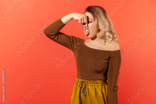 Portrait of blonde woman pinching nose, stop breathing bad odor, disgusted by smell of farting, her grimace expressing repulsion, gross. Stinky. Indoor studio shot isolated on red background