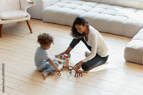 Long time ago... Smiling millennial african mother, elder sister or babysitter playing with small interested black boy on heated wooden floor with toy dinosaur figures and cubes creating fantasy world