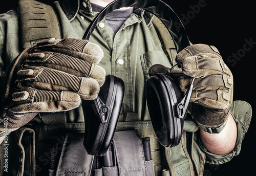 Soldier in tactical outfit with body armor, gloves and shirt holding shooting earmuffs on black background.