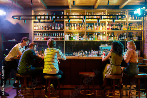 Rear view of three guys drinking beer, looking at women, two girlfriends sitting at the bar counter. Friends spending time at night club, restaurant