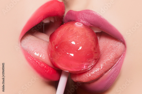 Sensual female mouth lick. Lesbian oral pleasure. Closeup view of two sexy sensual bright female lips with pink and purple lipgloss holding in mouth and licking delicious round red lollipop candy.