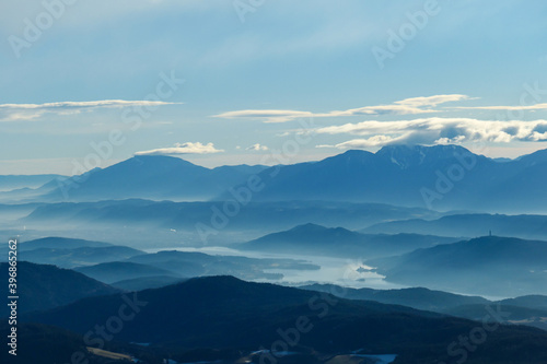 A panoramic view on Austrian Alps, captured on an early morning. The high peaks are hiding behind light clouds. The valley is shrouded in fog. There is a long lake at the bottom of the valley. Remedy