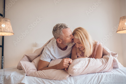 Grey-haired man and a blonde woman lying on the bed and looking romantic