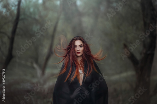 Mysterious fantasy gothic woman dark witch obsessed by evil. Red-haired Girl demon in black dress cape hood. Red hair flutters in wind. Dark dense deep forest background, trees. Scleral lenses on eyes