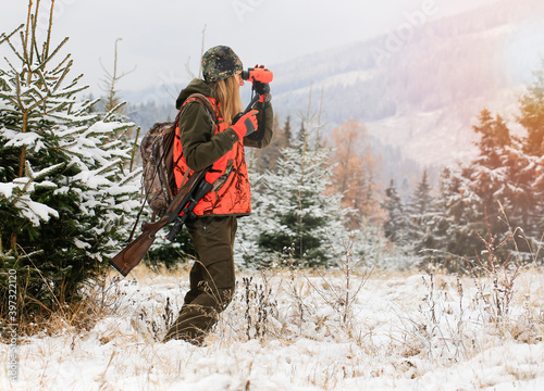 A young beautiful woman looks into the binocular with rifle on the shoulder ready for the hunt. There are snowy trees all around.