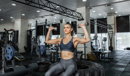 Muscular woman training shoulders doing dumbbell press sitting on cross wheel in a modern gym