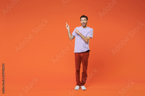 Full length of amazed excited surprised young bearded man 20s in basic violet t-shirt standing pointing index fingers up on mock up copy space isolated on bright orange background studio portrait.