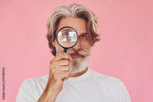 Using magnifying glass. Stylish modern senior man with gray hair and beard is indoors