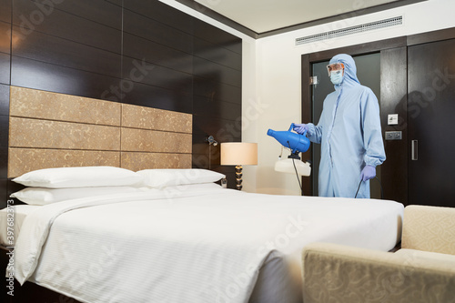 Worker in a special overalls standing in bedroom in hotel while disinfecting